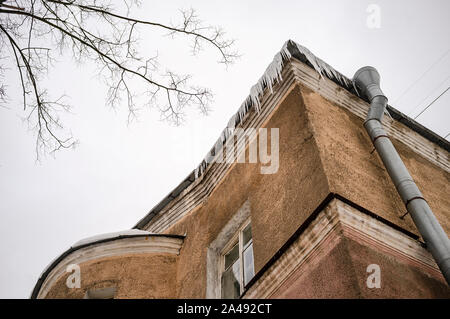 Les glaçons accrocher dangereusement sur le toit d'une maison à deux étages avec une gouttière et un arbre contre le ciel Banque D'Images