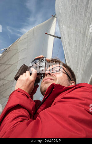 Un bel homme - un photographe en une veste rouge prend des photos sur un yacht, dans le contexte des voiles blanches et bleu ciel, sur une journée ensoleillée. Banque D'Images