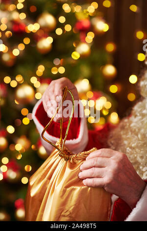 Le Père Noël ouvre un sac de cadeaux, assis près de l'arbre de Noël décoré avec des lumières - Joyeux Noël et bonnes fêtes ! Banque D'Images