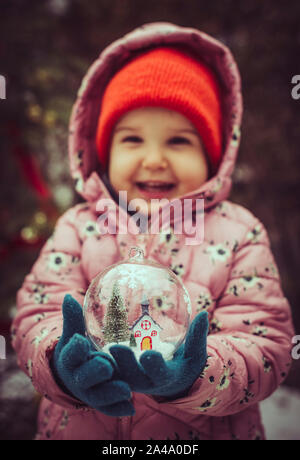 Petite fille tenir transparent bille de verre avec arbre de Noël et de l'intérieur de la chambre. La neige en boule de noël. Banque D'Images