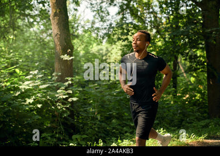 Homme africain avec corps sportif s'exécutant dans un jardin en matin d'été. Le port sportif en noir T-shirt et short à l'écoute de la musique avec des écouteurs. La formation de canaux chauds. Banque D'Images