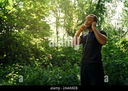 Beau runner en noir T-shirt et short noir debout dans la forêt et à l'écoute de la musique avec des écouteurs. Homme avec corps sportif posant avec les yeux fermés à l'extérieur. Banque D'Images