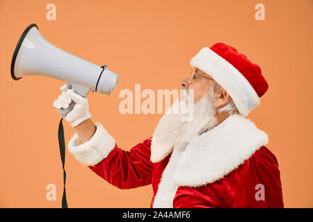 Vue latérale du Père Noël senior en rouge costume, gants blancs et les lunettes en parlant avec porte-voix pour annoncer à l'occasion de Noël. Genre grand-père posant en studio, avec un fond orange. Banque D'Images