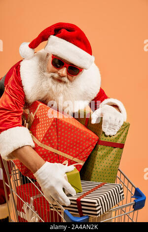 Cheerful man en costume traditionnel rouge, lunettes et gants blancs s'appuyant sur panier rempli de cadeaux colorés. Isolé sur fond orange. Studio shot. Banque D'Images