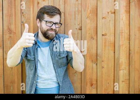 Un homme barbu, heureux de montrer les gestes verres Thumbs up sur deux mains et regarde dans la caméra, dans le contexte d'un vieux mur en bois. Close-up Banque D'Images