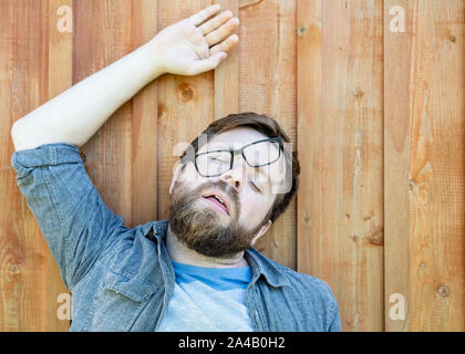 Homme barbu à lunettes était fatigué, de se reposer sur le plancher en bois et s'endormi avec sa bouche ouverte, dans une drôle de pose, à l'extérieur. Isolées. Fermer Banque D'Images