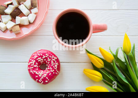 Une tasse de café, beignes et savoureux morceaux de noix de coco sur la table en bois blanc. Tulipe jaune fleurs, vue d'en haut. Plaque de couleur corail vivant et tasse. Banque D'Images