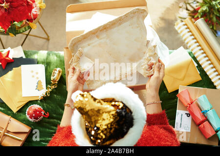 Saison de vacances. Vue de dessus de la femme moderne dans sparkle gold sequin santa hat got colis avec broken dish, assis sur la table dans le salon moderne un Banque D'Images