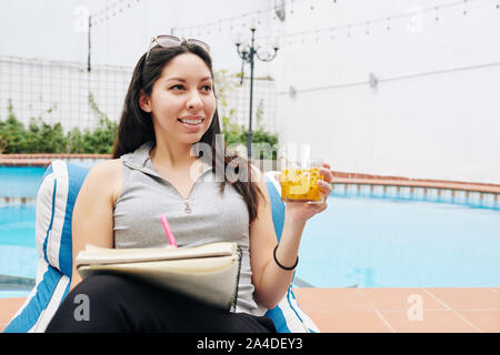 Portrait de jolie femme positive par repos piscine avec verre de jus de fruit et prendre des notes dans le journal Banque D'Images
