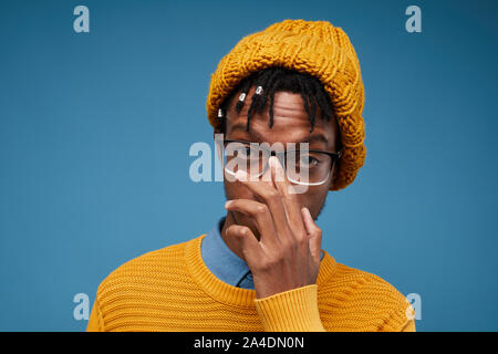 Portrait d'Afro-américain à la mode homme portant chapeau tricoté chandail lumineux et posant contre fond bleu hid réglage lunettes, copy space Banque D'Images