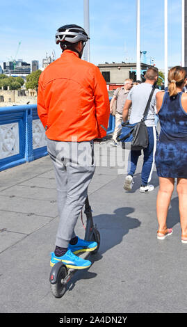 Vue arrière d'un homme portant un casque équitation Xiaomi Scooter électrique sur la chaussée derrière les piétons touristiques lentement le Tower Bridge Road London England UK Banque D'Images