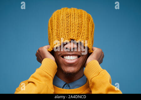 Portrait de vives émotions African man smiling at camera tout en tirant sur le jaune Knit hat posing contre fond bleu, copy space Banque D'Images