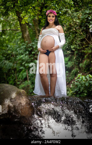 Belle femme enceinte latine 22-26 ans wearing white dress avec ventre montrant debout dans creek/stream Banque D'Images