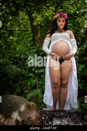 Belle femme enceinte latine 22-26 ans wearing white dress avec ventre montrant debout dans creek/stream Banque D'Images