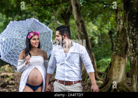 Happy young hispanic couple en train de marcher à l'extérieur. Une femme enceinte avec un parapluie et son mari profitez d'une promenade Banque D'Images