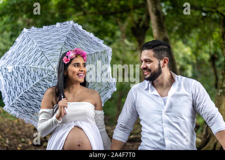 Happy young hispanic couple en train de marcher à l'extérieur. Une femme enceinte avec un parapluie et son mari profitez d'une promenade Banque D'Images