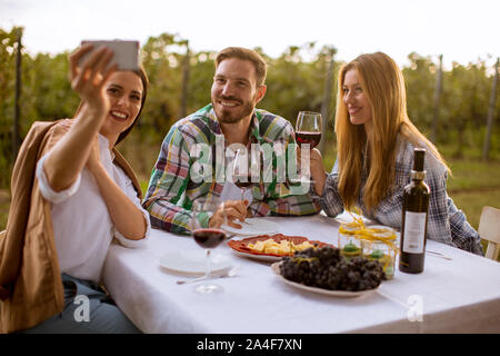 Groupe de jeunes gens assis près de la table, de boire du vin rouge et en tenant avec selfies téléphone mobile dans le vignoble Banque D'Images