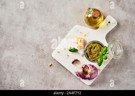 La sauce pesto genovese pesto ou dans un bocal en verre avec les noix de pin, le parmesan, le basilic, l'huile et l'ail sur la planche à découper en marbre blanc sur fond noir en gris. Banque D'Images