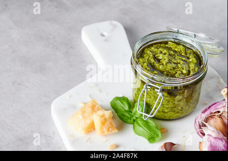 La sauce pesto genovese pesto ou dans un bocal en verre avec les noix de pin, le parmesan, le basilic, l'huile et l'ail sur la planche à découper en marbre blanc. Copier l'espace. Banque D'Images