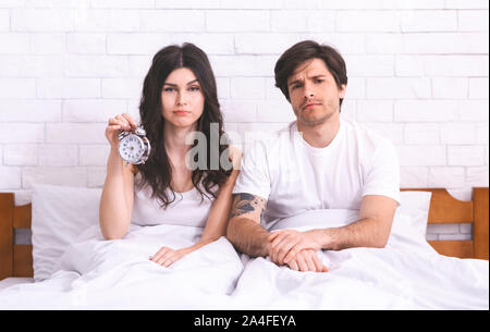 Sleepy couple sitting in bed avec réveil au matin Banque D'Images