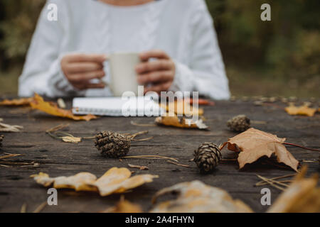 Jeune femme dans un pull blanc est assis à une vieille table sombre dans le parc, est titulaire d'une tasse de thé avec du café et lit et écrit sur l'arrière-plan de jaune Banque D'Images