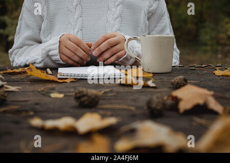 Jeune femme dans un pull blanc est assis à une vieille table sombre dans le parc, est titulaire d'une tasse de thé avec du café et lit et écrit sur l'arrière-plan de jaune Banque D'Images