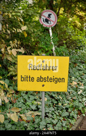 Nuremberg, Allemagne - 10 octobre 2019 : un panneau jaune dit Radfahrer bitte absteigen au début d'un jardin. Dans l'arrière-plan est un signe fo Banque D'Images