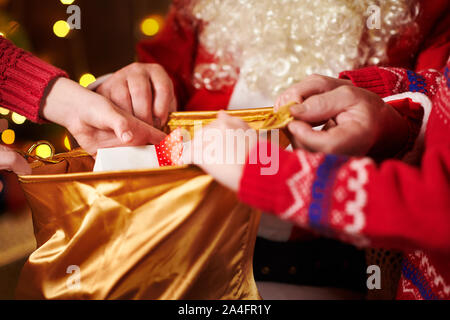 Le Père Noël ouvre un sac et donne des cadeaux aux enfants, assis près de l'arbre de Noël décoré avec des lumières - Joyeux Noël et bonnes fêtes ! Banque D'Images