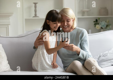 Heureux grand-mère mature de câlins petite-fille, à l'aide d'ensemble du smartphone. Banque D'Images