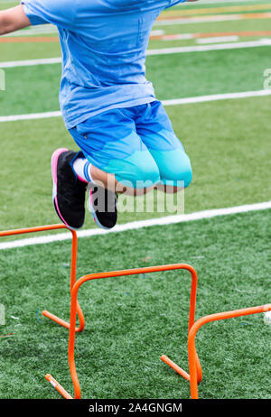 Un jeune garçon est sautant par-dessus un obstacle mini en plastique orange sur un champ de gazon vert au camp d'athlétisme. Banque D'Images