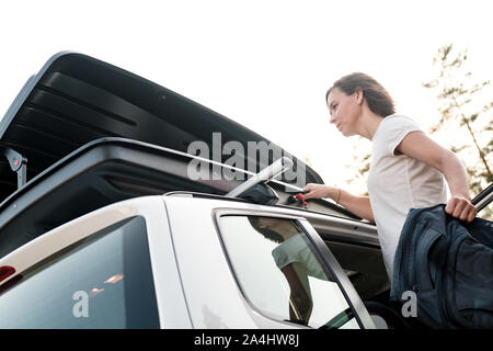 Une femme met les choses dans le toit d'une voiture ou dans une boîte de chargement, avant un voyage en famille en vacances, contre le ciel et les arbres, un soir d'été. Clo Banque D'Images