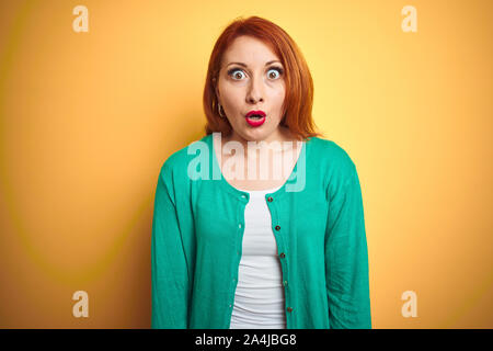 Belle jeune femme rousse portant chandail vert d'hiver isolé sur fond jaune peur et choqué par l'expression de surprise, la peur et l'excite Banque D'Images