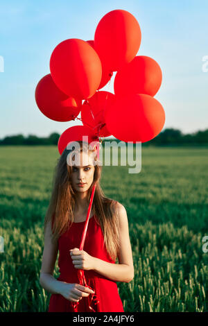Young Beautiful woman in red dress posing in green field avec des ballons rouges. La liberté, les loisirs, vacances Banque D'Images