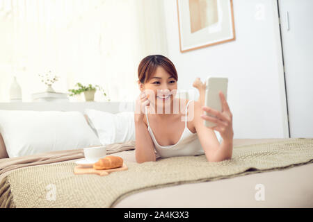 Young Asian woman lying on bed et l'utilisation de téléphone mobile, à son domicile. selfies Banque D'Images