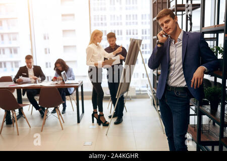 Young attractive homme élégant costume bleu à la consultation des clients par smartphone pendant que ses collègues travaillant sur l'atmosphère de travail du projet. Banque D'Images