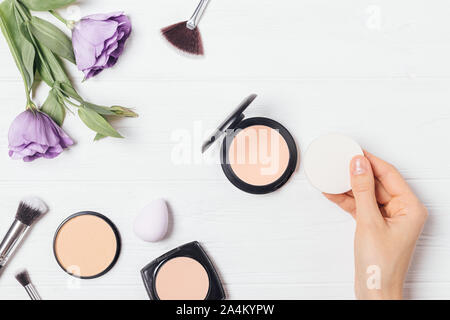 Vue supérieure de la femme main tenant à côté de l'éponge la poudre compacte parmi les produits et accessoires de maquillage, télévision floral composition laïcs. Banque D'Images