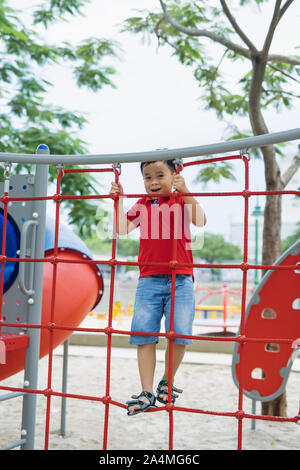 Jeune garçon asiatique monter sur la corde rouge clôture et barre grise de sa main, d'exercer à l'aire de porte de sortie sous le grand arbre. Banque D'Images