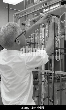 Années 1960, historique, un étudiant d'université de sexe masculin travaillant avec un ensemble de tubes à essai en verre fixé sur un cadre métallique dans un laboratoire de sciences, USA. Banque D'Images