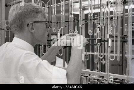 Années 1960, historique, un étudiant d'université de sexe masculin travaillant avec un ensemble de tubes à essai en verre fixé sur un cadre métallique dans un laboratoire de sciences, USA. Banque D'Images