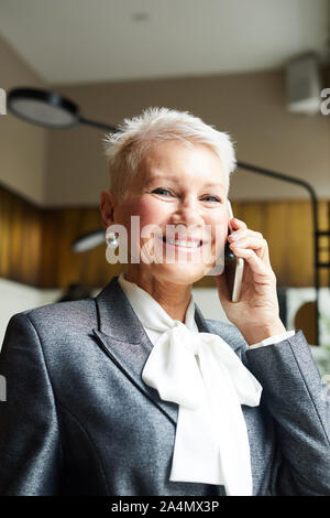 Portrait of mature businesswoman avec de courts cheveux blonds smiling at camera while talking on mobile phone at office Banque D'Images