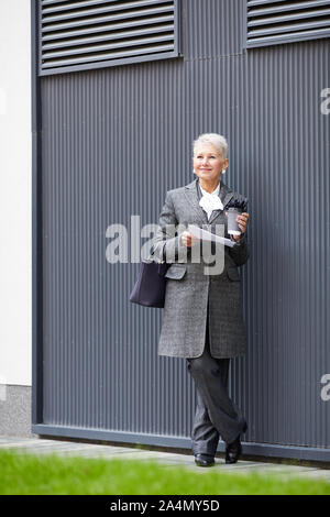 Femme mature blonde élégante avec des cheveux courts holding documents et boire du café dans ses mains tout en se tenant à l'extérieur Banque D'Images