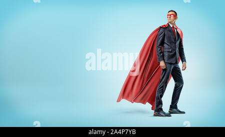 Un homme d'affaires dans un fluide en super-héros rouge cape et un masque regardant par-dessus son épaule sur fond bleu. Banque D'Images