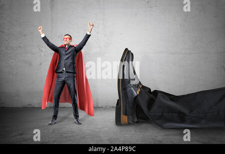 A smiling businessman dans une cape rouge de super-héros avec les mains posées dans la réussite motion debout à côté d'un pied humain géant. Banque D'Images