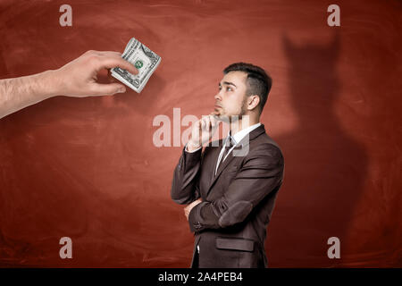 Une main géante avec un rouleau de billets de $1 en face d'une pensée d'affaires qui font de l'ombre d'un diable. Banque D'Images