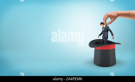 Un petit homme d'affaires sur un fond bleu tiré d'un chapeau d'illusionniste par un grand homme de main. Banque D'Images