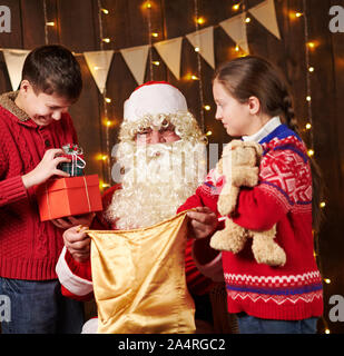 Le Père Noël ouvre un sac et donne des cadeaux aux enfants, assis près de l'arbre de Noël décoré avec des lumières - Joyeux Noël et bonnes fêtes ! Banque D'Images