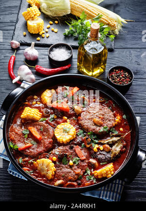 Les burgers de viande en sauce marinara, avec des champignons, des légumes et des cors dans une cocotte en céramique noire avec des ingrédients sur une pierre noire sur un woode Banque D'Images