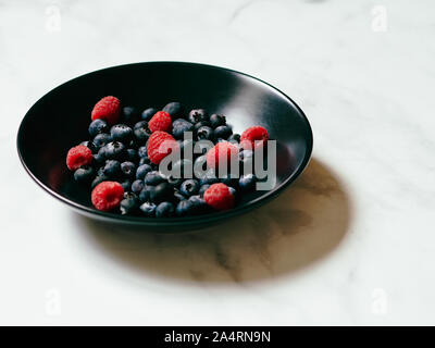 Les bleuets et les framboises dans un plat noir sur un fond de marbre blanc en studio Banque D'Images