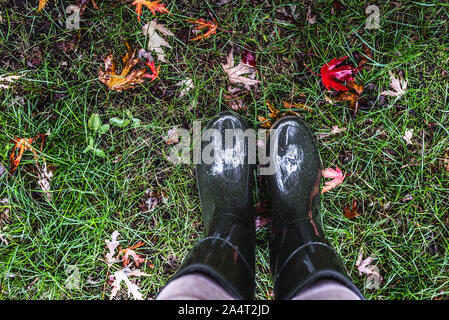 Les pieds dans des bottes en caoutchouc vert olive debout sur l'herbe verte avec des feuilles mortes. Banque D'Images