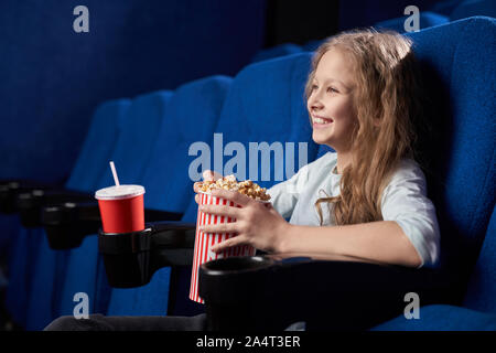 Vue latérale d'une joyeuse fille rousse riant de comédie au cinéma. Heureux enfant femelle eating popcorn, boire de l'eau douce de la relaxine et aérés Banque D'Images